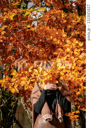 A woman stands near an autumn tree, covering her face with a colorful fall leaf, mid shot, outdoors, seasonal mood A woman stands near an autumn tree, covering her face with a colorful fall leaf, mid shot, outdoors, seasonal mood 132885827