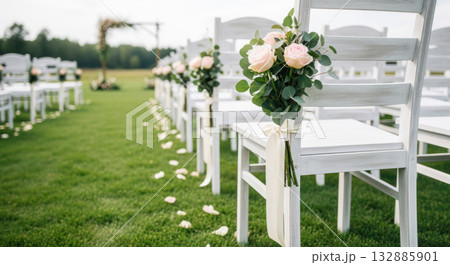 Elegant outdoor wedding ceremony setup featuring white chairs adorned with floral arrangements and scattered rose petals on lush green grass Elegant outdoor wedding ceremony setup featuring white chairs adorned with floral arrangements and scattered rose petals on lush green grass 132885901