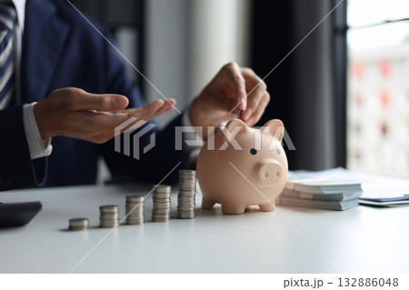 Businessman in a suit putting a coin into a piggy bank with stacks of coins and money on the table. Concept of saving money and financial planning, financial growth, investment, and future planning. 132886048