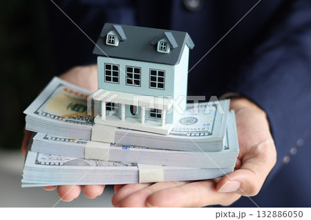 Businessman holding a house model on top of stacks of money, representation of real estate investment, home loan, selling, renting, insurance. Businessman holding a house model on top of stacks of money, representation of real estate investment, home loan, selling, renting, insurance. 132886050