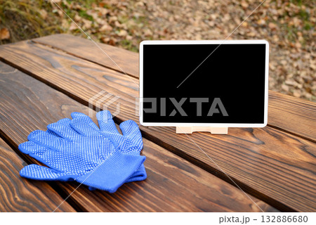 a blue gardening glove and a white tablet on a stand are on a brown wooden table 132886680