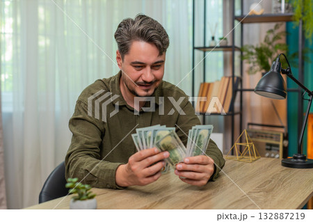 Middle-aged man counts cash smiling showing big financial success dollar bills wealth at table desk 132887219
