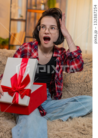 Young happy woman opening gift at home with surprise and joy, reacting with happiness, wide smile 132887254
