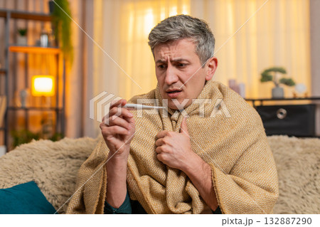 Young man checks body temperature with thermometer looks worried about possible illness and fever Young man checks body temperature with thermometer looks worried about possible illness and fever 132887290
