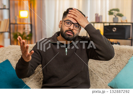 Indian man on sofa at home covers face with hand in frustration, reacting with facepalm gesture Indian man on sofa at home covers face with hand in frustration, reacting with facepalm gesture 132887292