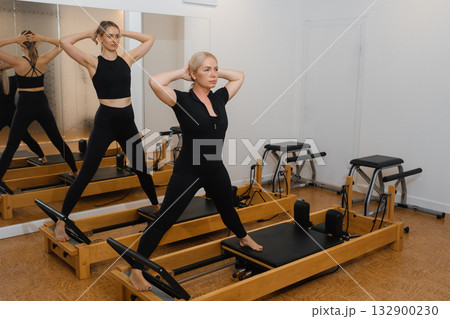 Two women engage in a Pilates workout at a modern studio focusing on core strength and flexibility in a bright, mirrored space in the morning hours Two women engage in a Pilates workout at a modern studio focusing on core strength and flexibility in a bright, mirrored space in the morning hours 132900230