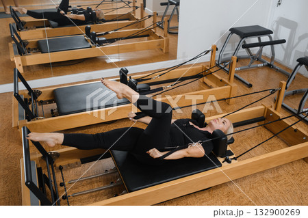 Woman practicing Pilates on a reformer machine in a well-lit studio with wooden floors and mirrors during a workout session Woman practicing Pilates on a reformer machine in a well-lit studio with wooden floors and mirrors during a workout session 132900269