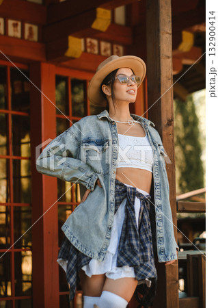 Stylish young woman poses confidently in a sunlit outdoor setting wearing denim, a hat, and sunglasses during a warm afternoon Stylish young woman poses confidently in a sunlit outdoor setting wearing denim, a hat, and sunglasses during a warm afternoon 132900414