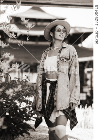Stylish woman in denim jacket and hat stands confidently in a vibrant outdoor setting with flowers and traditional architecture in the background during daylight 132900418