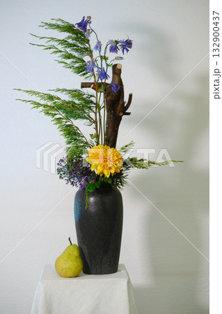 Elegant flower arrangement featuring yellow and purple blooms in a black vase with natural elements and a pear placed beside it on a white surface 132900437