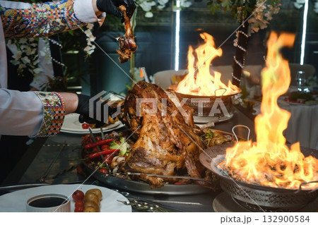Guests enjoy traditional feast with roasted meats and vibrant flames at festive gathering in elegant setting Guests enjoy traditional feast with roasted meats and vibrant flames at festive gathering in elegant setting 132900584