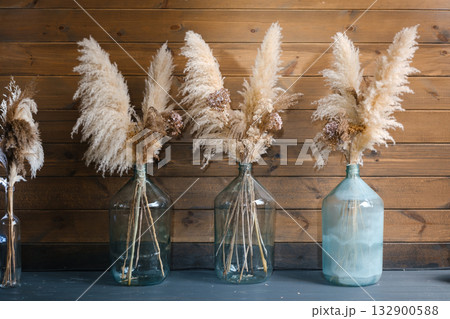 Dried pampas grass arranged in glass vases on a wooden table, creating a rustic and warm atmosphere in interior decor Dried pampas grass arranged in glass vases on a wooden table, creating a rustic and warm atmosphere in interior decor 132900588