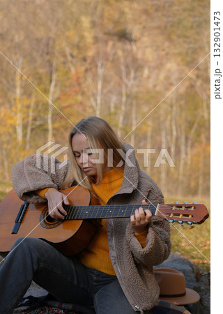 Beautiful blonde girl playing acoustic guitar and singing relaxing enjoying outdoors on sunny autumn day. Hippie woman in boho style learning and practicing playing musical instrument. Fall leaves Beautiful blonde girl playing acoustic guitar and singing relaxing enjoying outdoors on sunny autumn day. Hippie woman in boho style learning and practicing playing musical instrument. Fall leaves 132901473