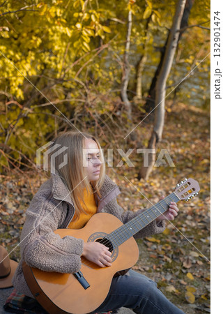 Blonde woman playing string guitar outdoors in autumn forest. Concept of sound therapy, mental health and wellness rituals. Calmness tranquility audio-sensory practices. Aura farming energy 132901474