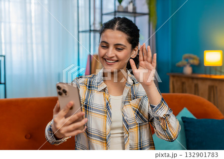 Indian woman on sofa happily waving hand greeting via video call on smartphone smiling joyful Indian woman on sofa happily waving hand greeting via video call on smartphone smiling joyful 132901783