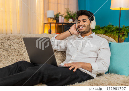 Indian man enjoying music, wearing wireless headphones, nodding head to rhythm, using laptop on sofa 132901797