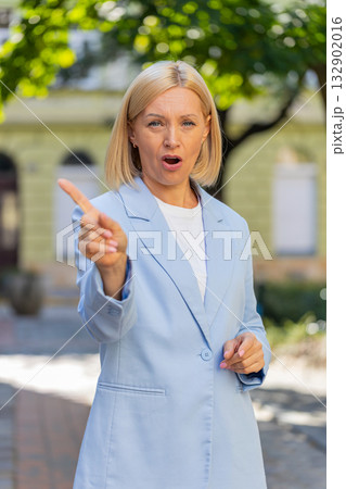 Caucasian mature businesswoman in suit standing in city street looking at camera shaking finger 132902016