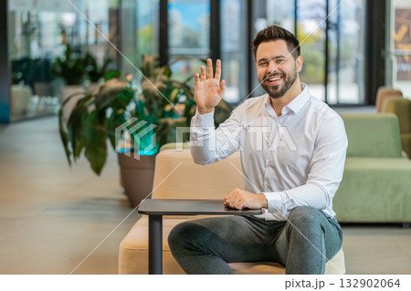 Portrait of smiling young businessman with laptop wave hand at camera sitting in hotel lobby 132902064