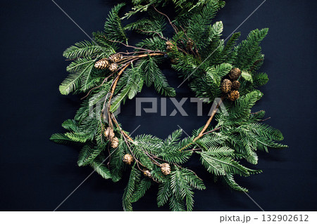 A minimalist holiday wreath made of evergreen branches, pinecones, and twigs on a dark background. Rustic elegance, natural textures, Scandinavian style, modern cabin trends 132902612