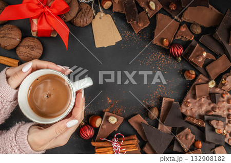 woman holding cup of hot chocolate surrounded by assorted dark and milk chocolate pieces, hazelnuts, cookies, Christmas decorations on dark background copy space Top view 132908182