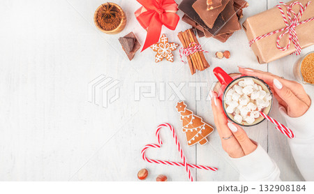 female hands holding red mug of hot chocolate with marshmallows and candy cane surrounded by gingerbread cookies, chocolate, cinnamon, and festive gifts on light background copy space. Top view 132908184