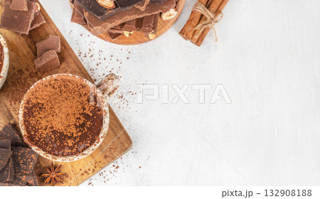 Top view of cup of hot chocolate sprinkled with cocoa powder on wooden board with cinnamon sticks, star anise, and chocolate pieces, cozy winter drink concept with copy space on light background 132908188