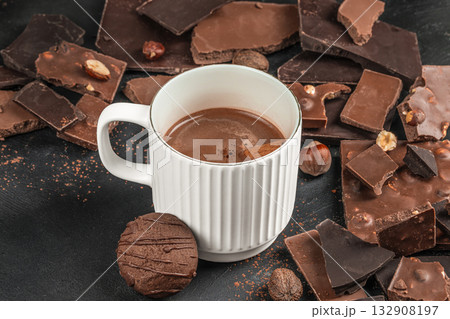 Close-up of white ceramic mug with hot chocolate and cookie surrounded by dark and milk chocolate pieces with hazelnuts on black textured background, cozy winter drink concept, top view copy space Close-up of white ceramic mug with hot chocolate and cookie surrounded by dark and milk chocolate pieces with hazelnuts on black textured background, cozy winter drink concept, top view copy space 132908197