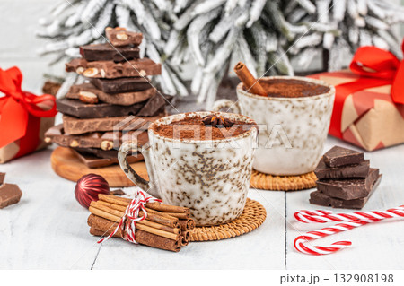 Two ceramic cups of hot chocolate with cinnamon sticks and anise on white table, surrounded by Christmas gifts, candy canes, chocolate pieces, cozy winter holiday festive atmosphere 132908198