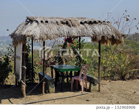 Rustic hut viewpoint A small thatched hut with wooden posts and plastic chairs overlooking lush green hills under clear skies, evoking rural tranquility. 132908906