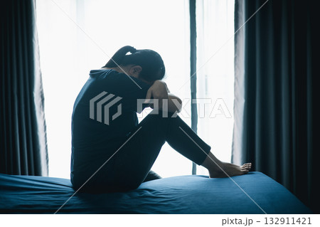 A stressed woman sitting alone on bed in dark room, holding her head down, symbolizing depression, anxiety, hopelessness, loneliness, mental health struggle, burnout and emotional pain. 132911421