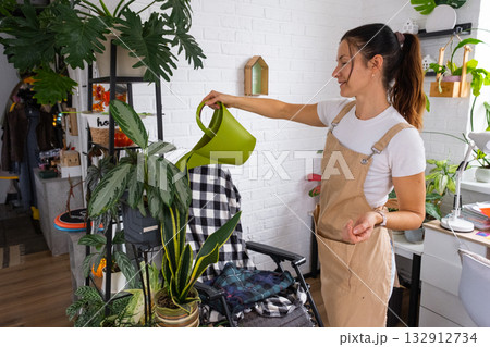 A woman waters home plants from her collection of rare species from a watering can, grown with love on shelves in the interior of the house. Home plant growing, green house, water balance 132912734