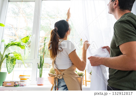 Husband helps his wife hangs transparent tulle curtains on large windows in the house inside the interior with potted plants. Spring cleaning, tidying up 132912817