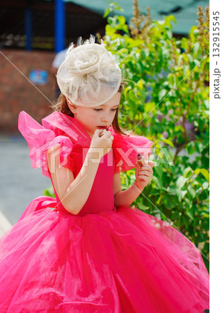 A cute little girl in a lush pink dress and hat Painting Her Lips. A cute little girl in a lush pink dress and hat Painting Her Lips. 132915545