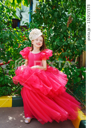 A girl in a lush pink dress and hat near the green bushes 132915578