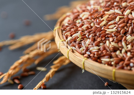Close up of organic brown rice grains in a bamboo basket with golden rice stalks on black stone background. 132915624
