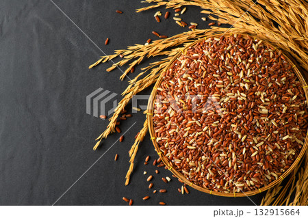 Close up of organic brown rice grains in a bamboo basket with golden rice stalks on black stone 132915664