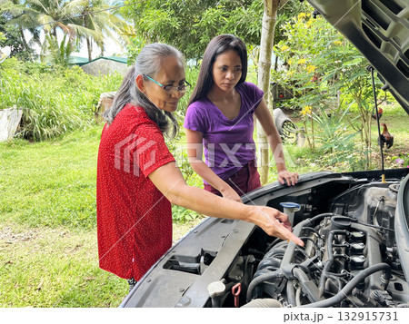 An elderly and young Asian woman looks under the hood of a broken down car to find a problem with the engine 132915731