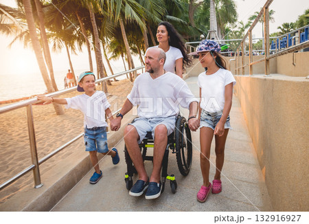 Disabled man in a wheelchair moves on a ramp to the beach with his family. Disabled man in a wheelchair moves on a ramp to the beach with his family. 132916927