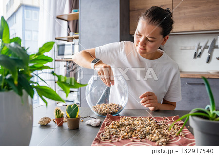 Young woman pouring stones into a glass container while preparing a terrarium. Concept of mindful crafting, eco-friendly lifestyle and home plant design. Young woman pouring stones into a glass container while preparing a terrarium. Concept of mindful crafting, eco-friendly lifestyle and home plant design. 132917540