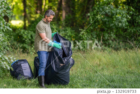 Man in rubber gloves with trash bag clean up garbage. Man collecting plastic. Eco volunteer. Garbage collecting in forest. Ecology concept. Volunteer collecting garbage, ecology. Social advertisement 132919733