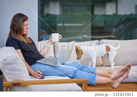 Woman with laptop and dog relaxing on terrace sofa with coffee 132921921