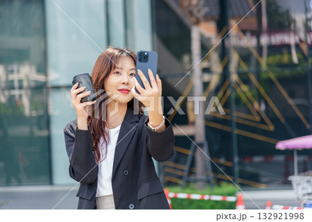 Young Asian woman business work suit on city street, busy taking phone selfie, holding coffee cup, bright sunlight highlights professional modern style, urban downtown scene visible. 132921998