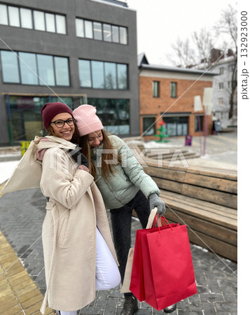 Two young women smiling outdoors with shopping bags in winter. Friendship, laughter, and lifestyle moments during city leisure and cold weather. Two young women smiling outdoors with shopping bags in winter. Friendship, laughter, and lifestyle moments during city leisure and cold weather. 132923000