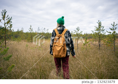Rearview of woman with backpack walk through marshy terrain among tall grass under overcast sky Rearview of woman with backpack walk through marshy terrain among tall grass under overcast sky 132923879