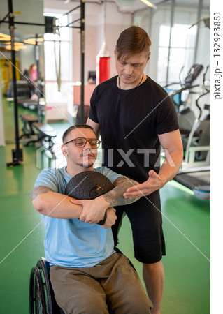 Fitness trainer assisting man in wheelchair during med ball workout on inclusive training in gym. 132923881
