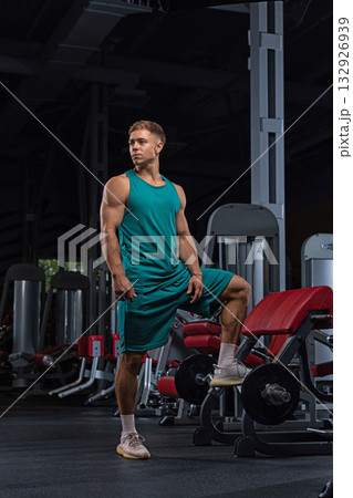 fit man stands in a gym one leg resting on gym equipment showcasing toned muscles. He appears focused and ready for his workout surrounded by exercise machines. 132926939