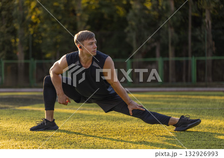 man is stretching on a football field. He wears headphones and focuses on his exercise under the warm sun. The grass is green and the surroundings are calm. man is stretching on a football field. He wears headphones and focuses on his exercise under the warm sun. The grass is green and the surroundings are calm. 132926993