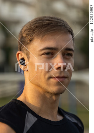 male athlete in a black sleeveless shirt listens to music with headphones while getting ready for an evening workout at a sports ground. The sun sets in the background. 132926997