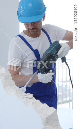 Male construction worker demolishing concrete wall with rotary hammer, wearing protective safety overalls is generating dust and debris during renovation project. Vertical portrait view Male construction worker demolishing concrete wall with rotary hammer, wearing protective safety overalls is generating dust and debris during renovation project. Vertical portrait view 132928109