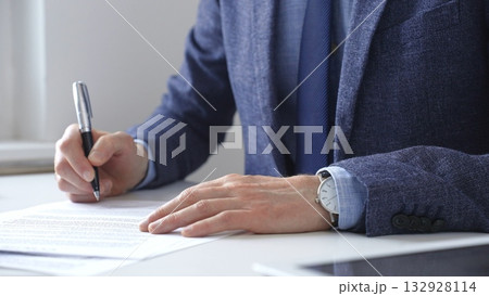Professional businessman signing contract with silver pen, wearing tailored blue suit, business documents and digital tablet nearby, close up of hands. Business people concept 132928114